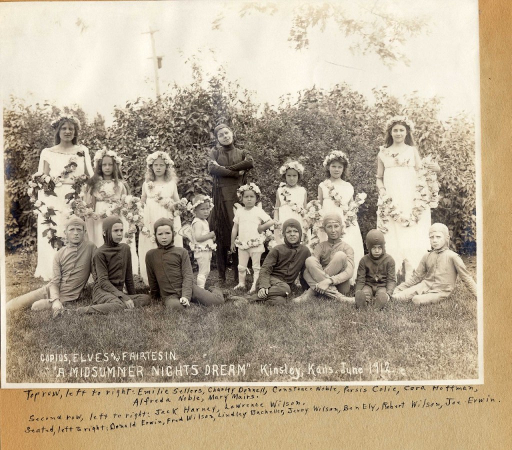 A vintage photograph featuring children in costumes for a performance of 'A Midsummer Night's Dream' in Kinsley, Kansas, June 1912. The group is arranged in two rows, with floral decorations and nature in the background.